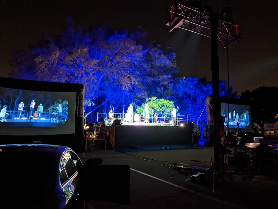Collin Theatre students and alumni perform on stage at Park-A-Palooza in the Plano Campus parking lot while spectators watch from their cars.