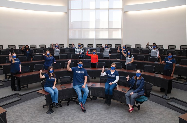Collin College students pose in a classroom while holding up their index fingers to represent the first class of baccalaureate graduates from the college.