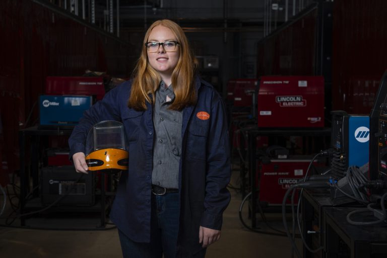 Picture of Cassie Dearing, Collin College welding student, holding a welding helmet and wearing safety glasses with the welding workshop in the background.