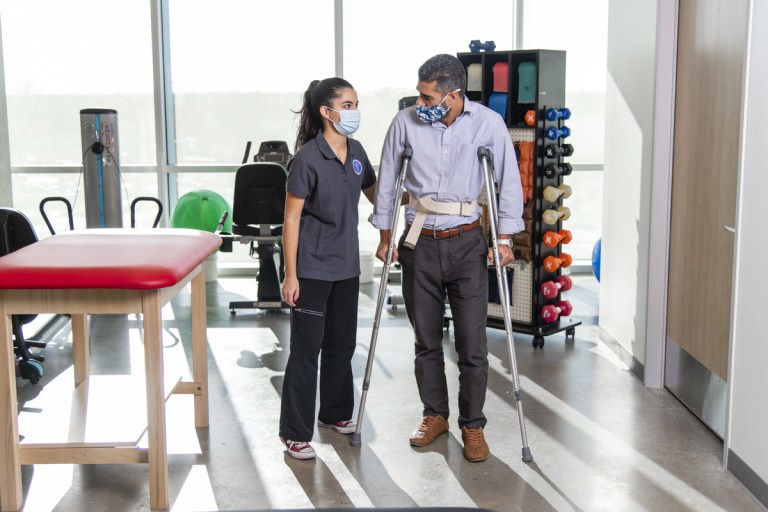 Rehabilitation Aide student supports a man on crutches.